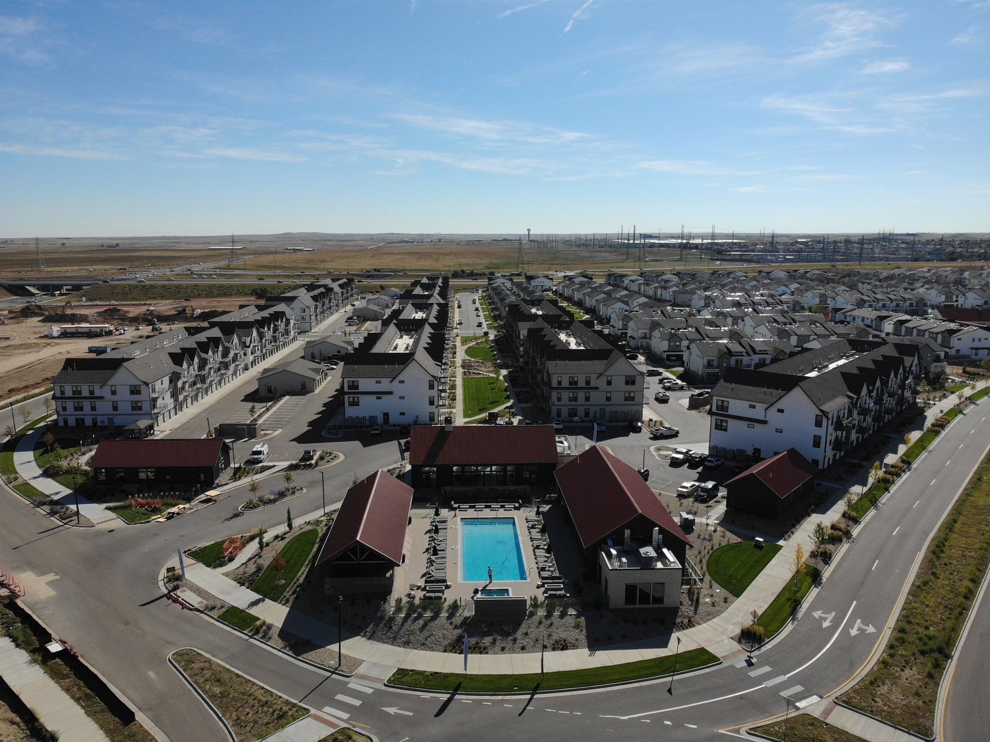 Aerial view of a modern suburban neighborhood with rows of houses, a central swimming pool, green spaces, and wide roads curving around the community under a clear blue sky.