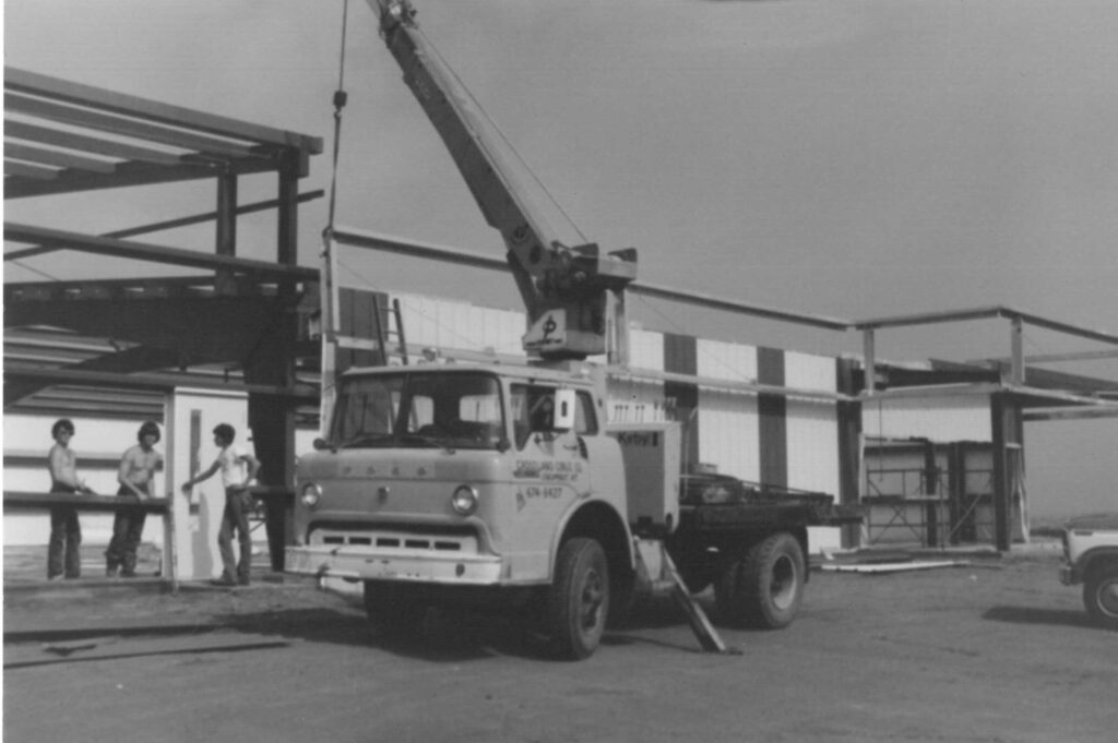 A black-and-white photo of a Ford crane truck on a construction site, with its boom raised. Four workers stand nearby, assembling the steel framework of a building under construction.