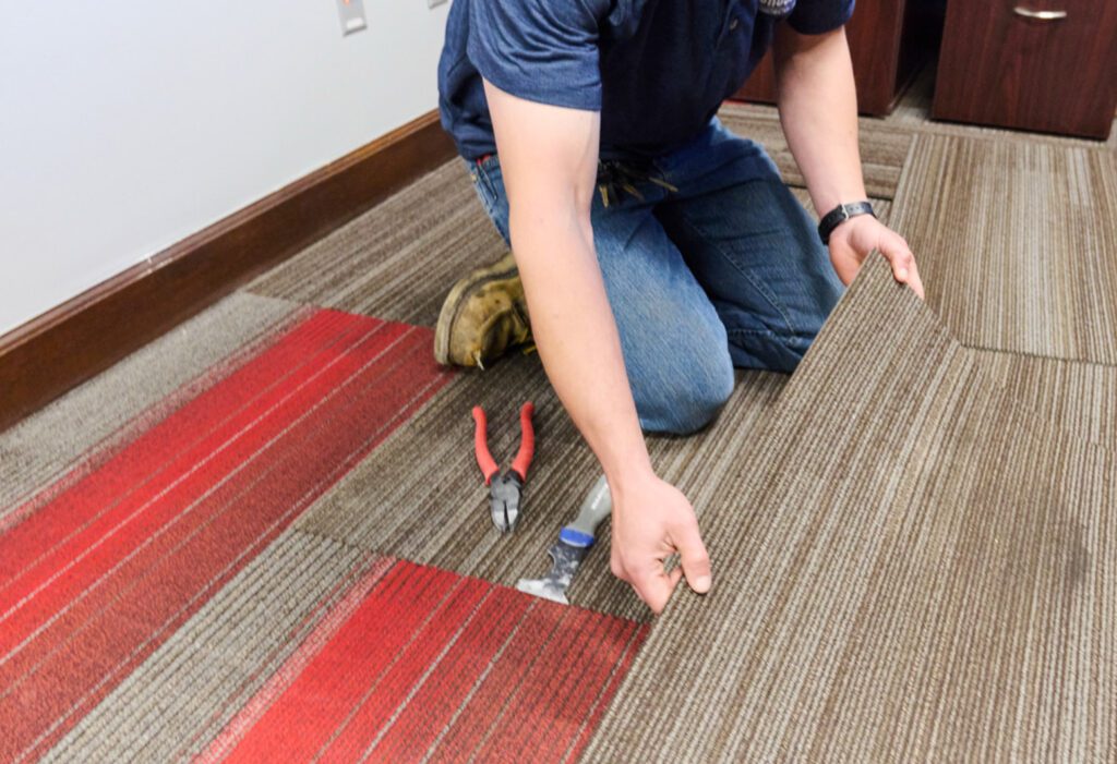 A person kneels on the floor, installing brown and red striped carpet tiles. Tools including pliers and a utility knife are nearby.