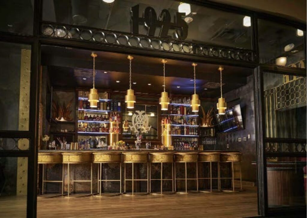 A stylish bar with gold accents, five gold pendant lights, and a row of gold barstools. Shelves behind the bar hold bottles and glassware, and “1925” is displayed above the bar in large numbers.