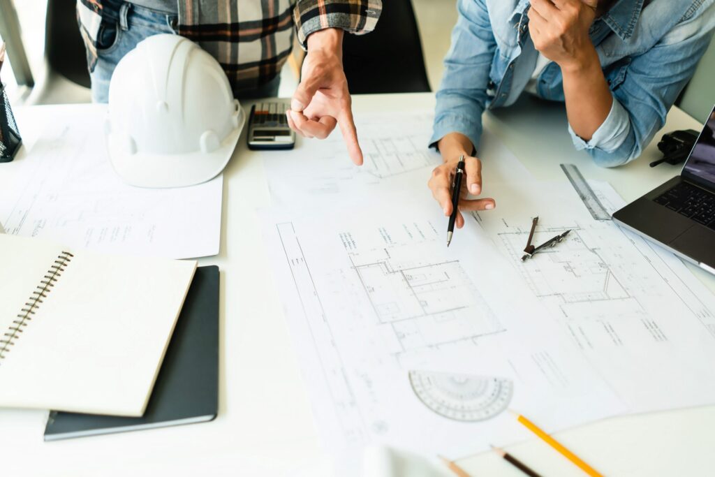 Two people pointing at a floor plan on a desk laid out with a hard hat, compass, scale rule and laptop.