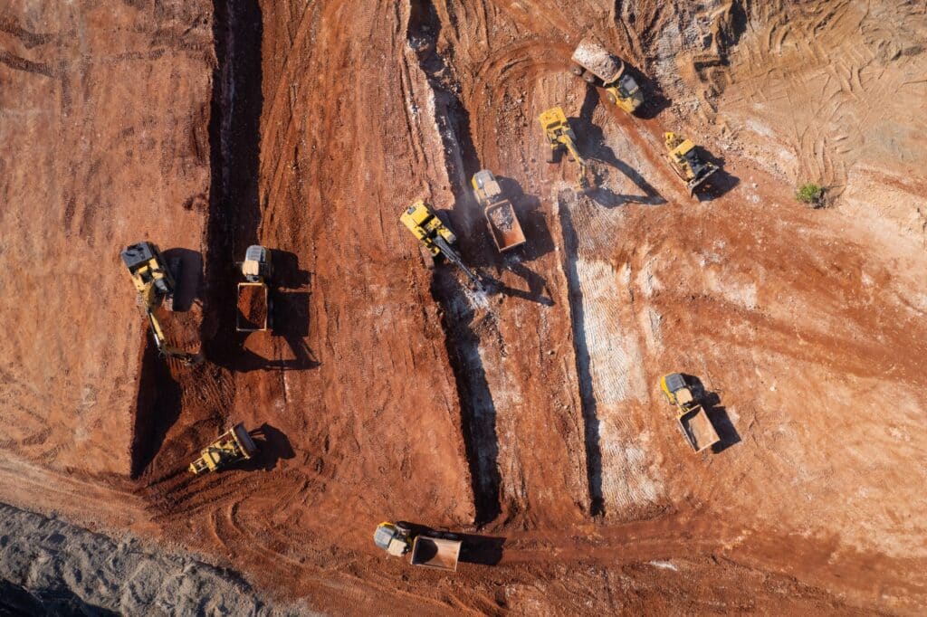 Aerial view of excavators and haul trucks cutting trenches across a red-dirt earthwork site.