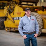 A man wearing a gray button-down shirt, jeans, and a cap stands smiling in front of large yellow construction equipment inside an industrial building.
