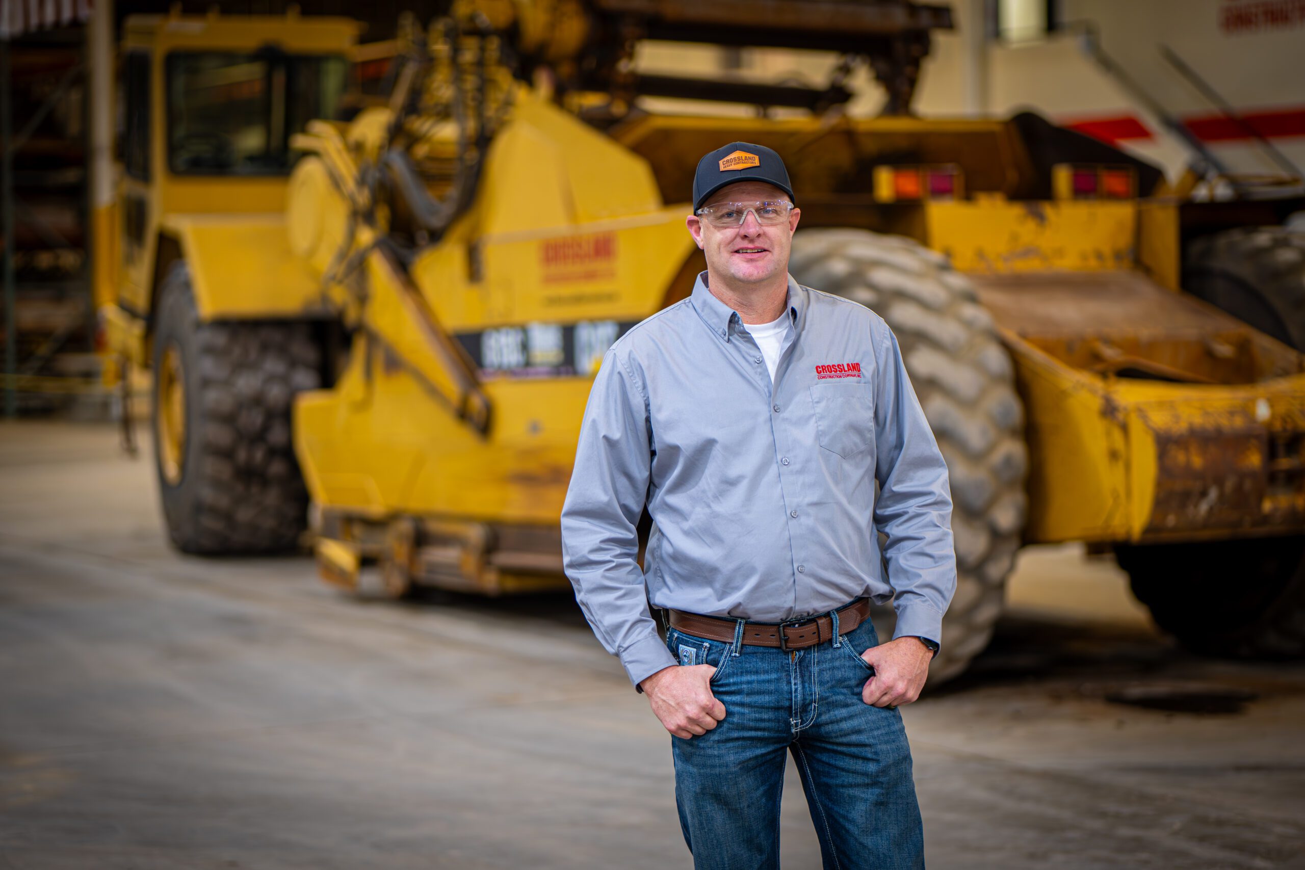 A man wearing a gray button-down shirt, jeans, and a cap stands smiling in front of large yellow construction equipment inside an industrial building.