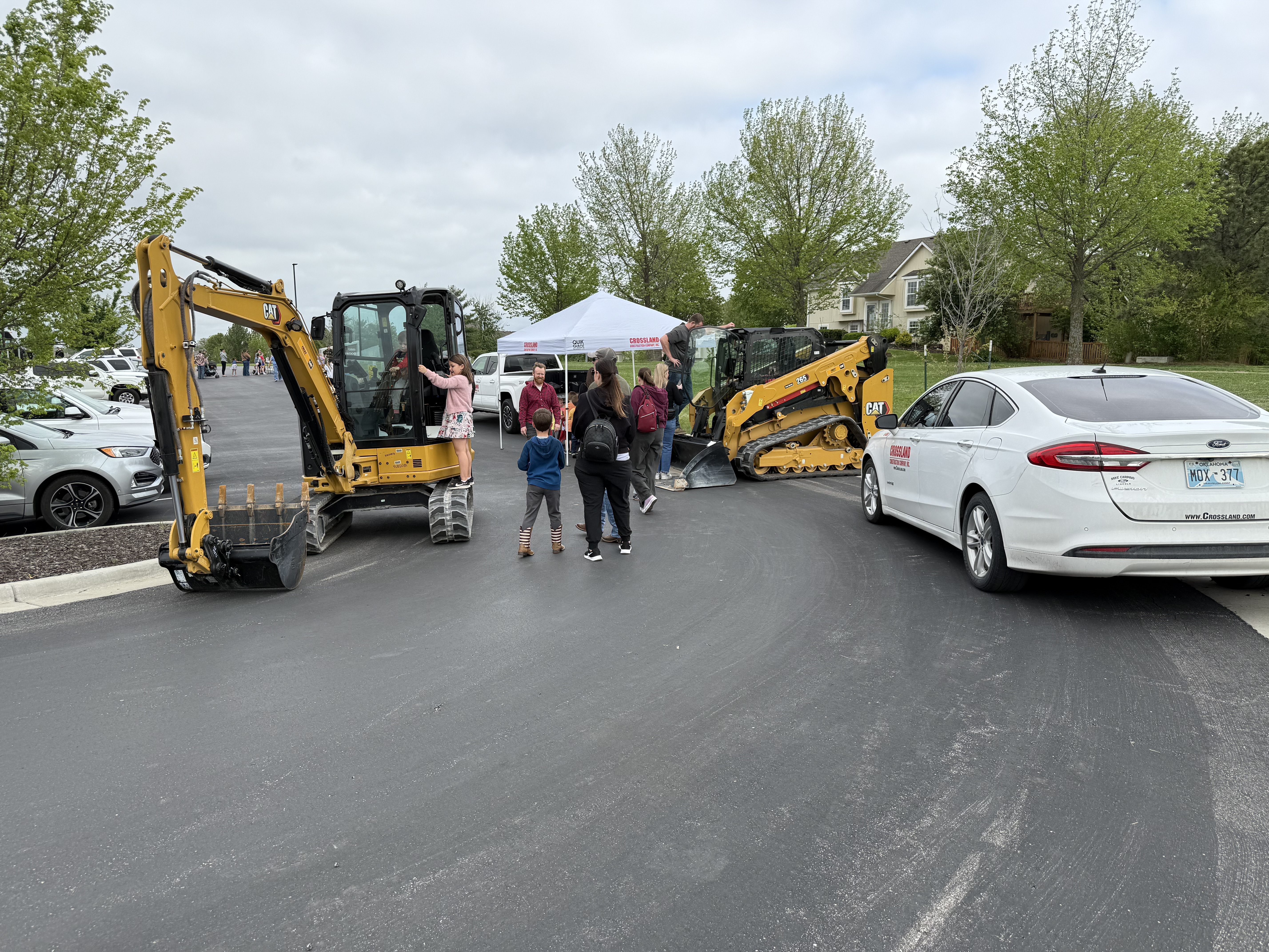 A group of people, including children, gather near two construction vehicles and a white tent in a parking lot; a white car is parked nearby, and trees and houses are visible in the background.