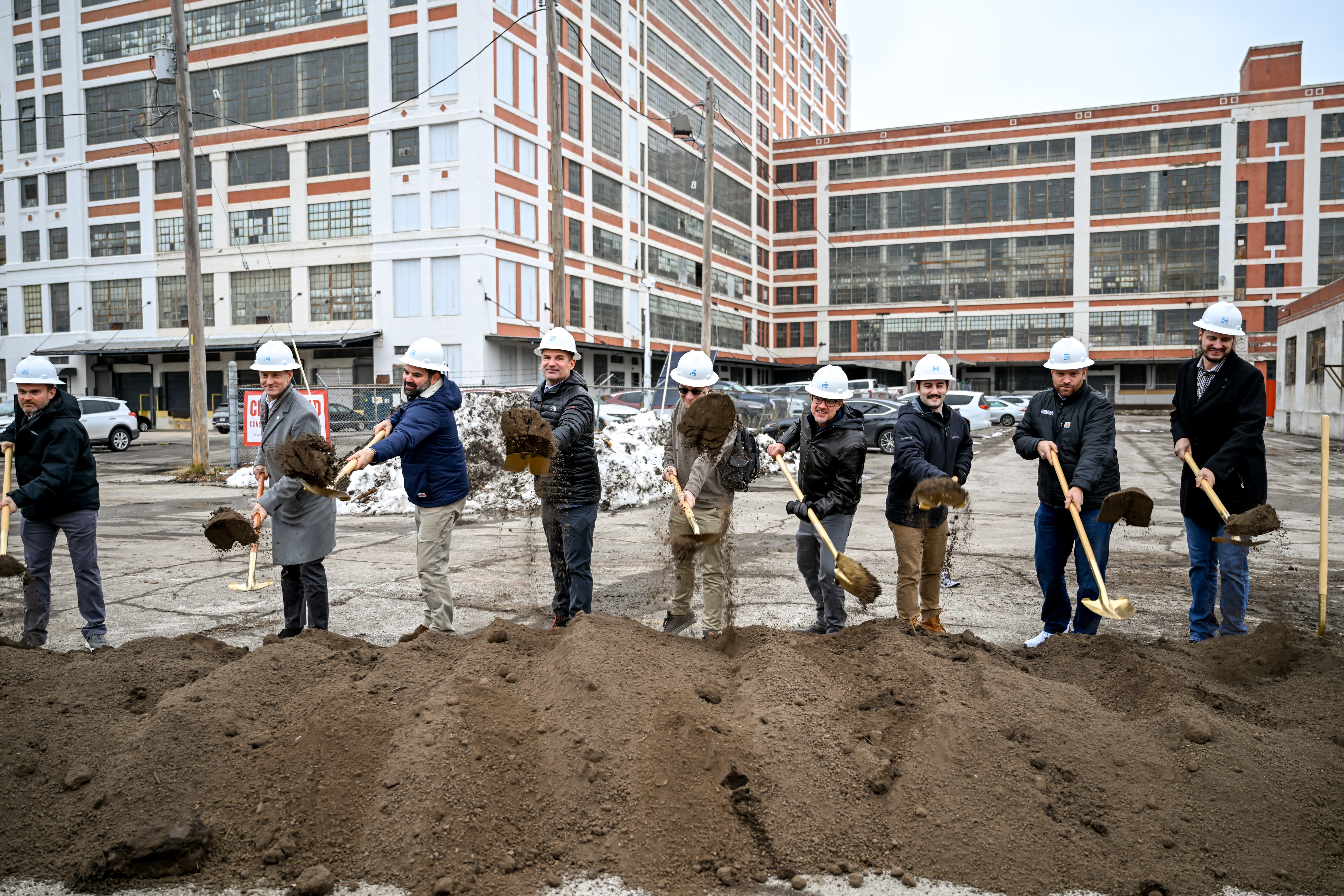 A group of people wearing hard hats and holding shovels participate in a groundbreaking ceremony, digging into a mound of dirt in front of a large industrial building.