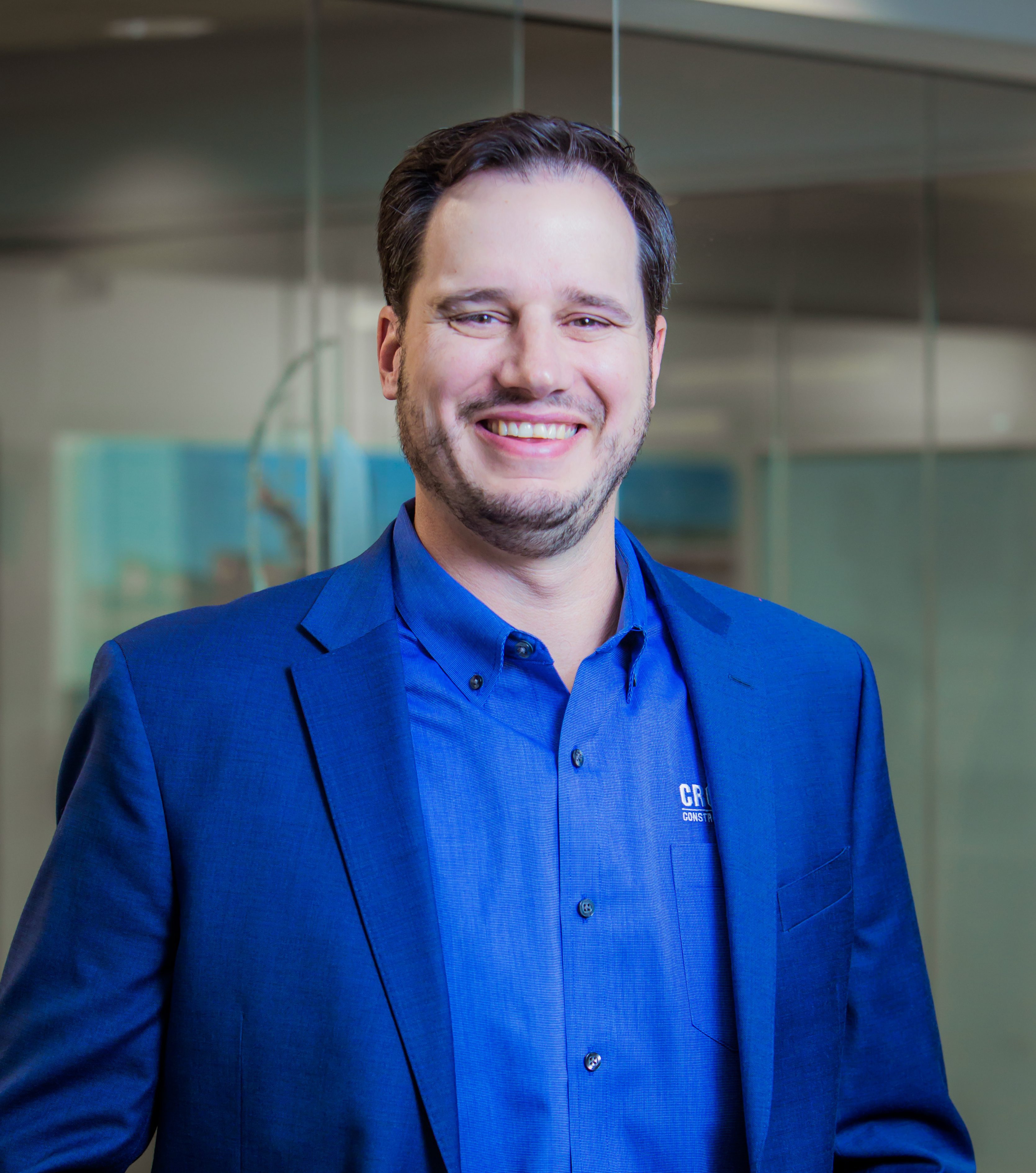 A man with short brown hair and a beard, wearing a blue blazer and blue shirt, smiles while standing in an office with glass walls.