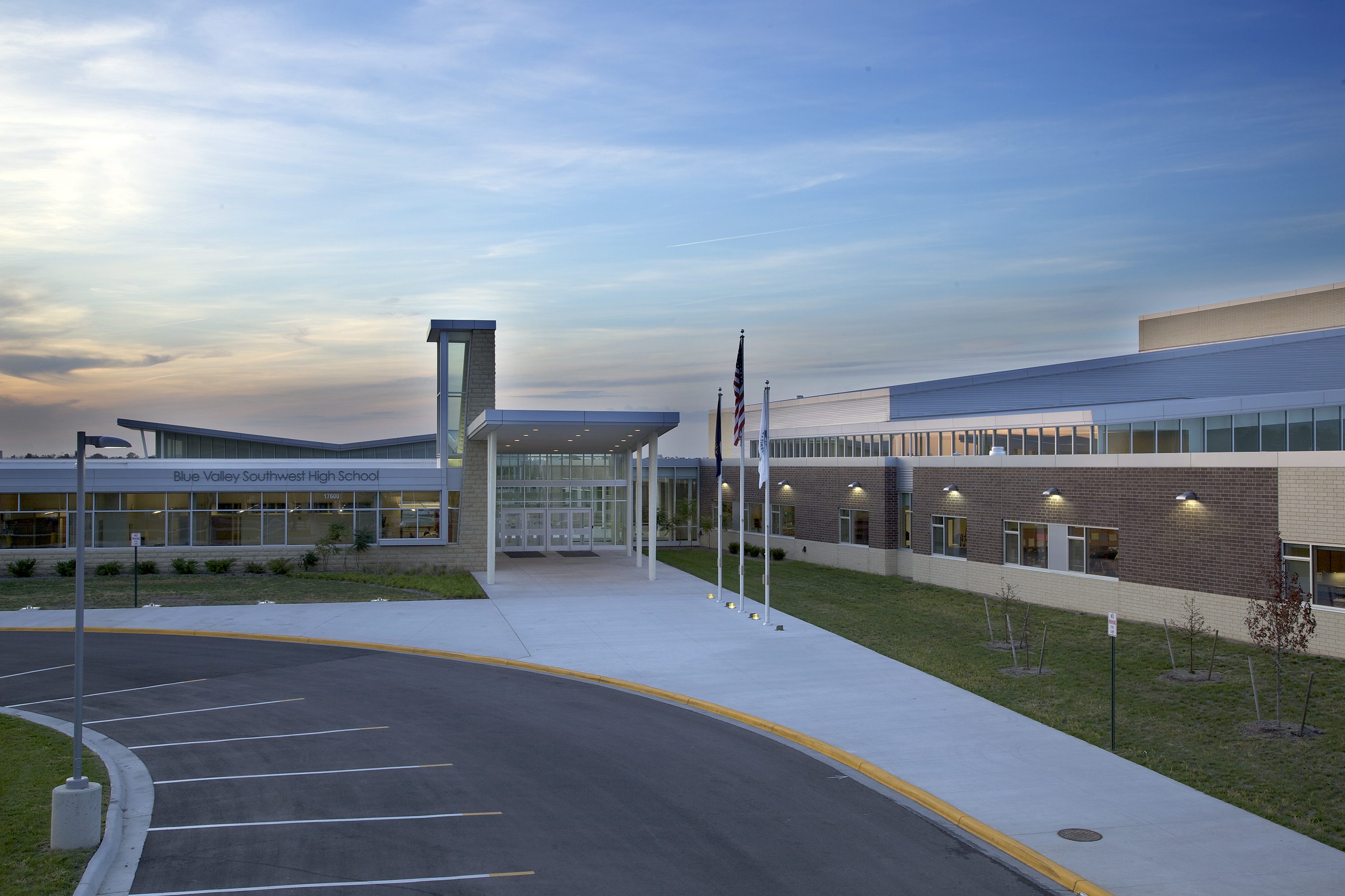 Modern high school building with large windows, brick and beige exterior, and a covered entrance. Three flagpoles stand in front, with a curved driveway and parking area. The sky is clear with a sunset glow.