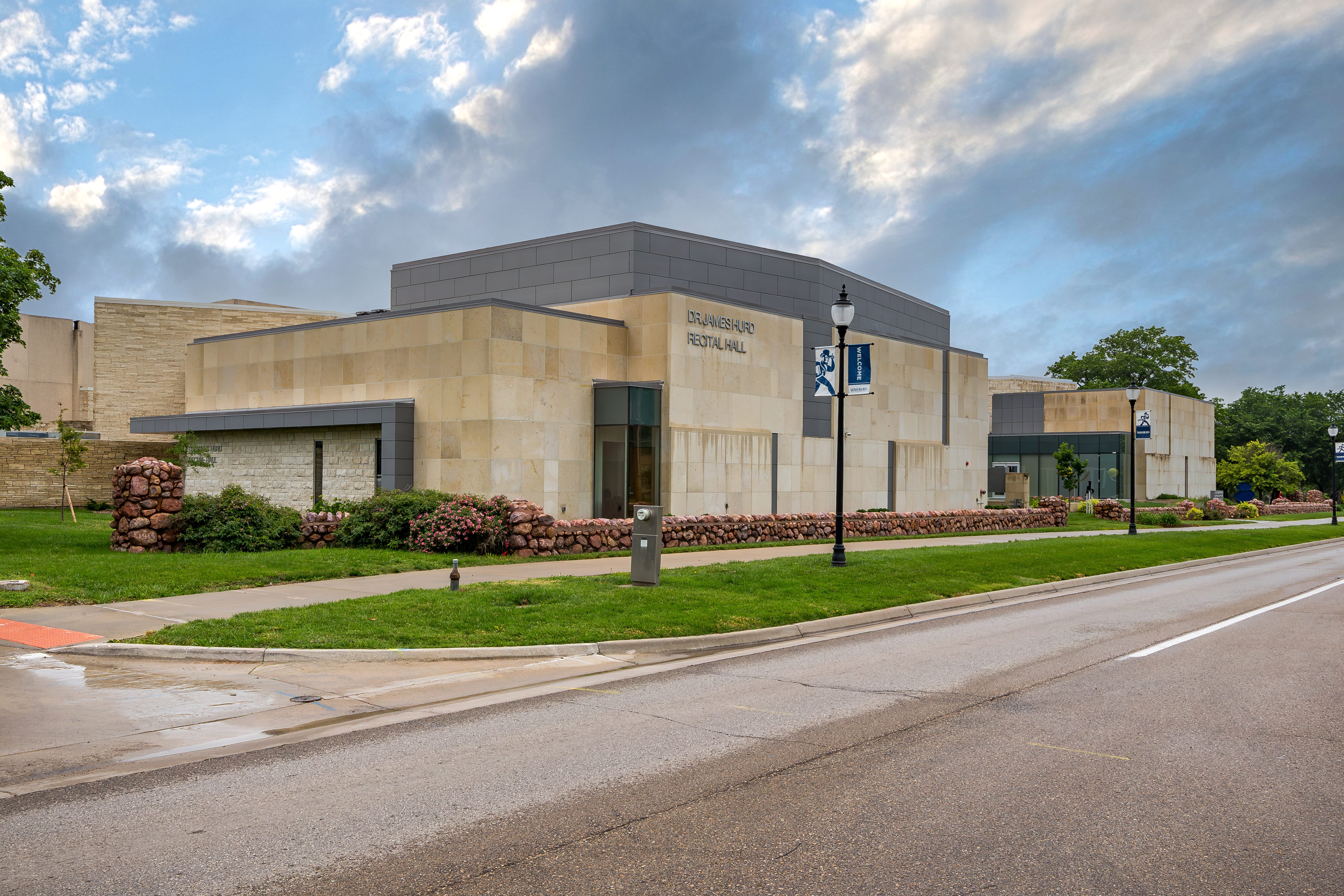 A modern, light stone and gray building labeled "Brannigan Recital Hall" sits beside a street, with green grass, landscaped bushes, and a few trees under a cloudy sky.