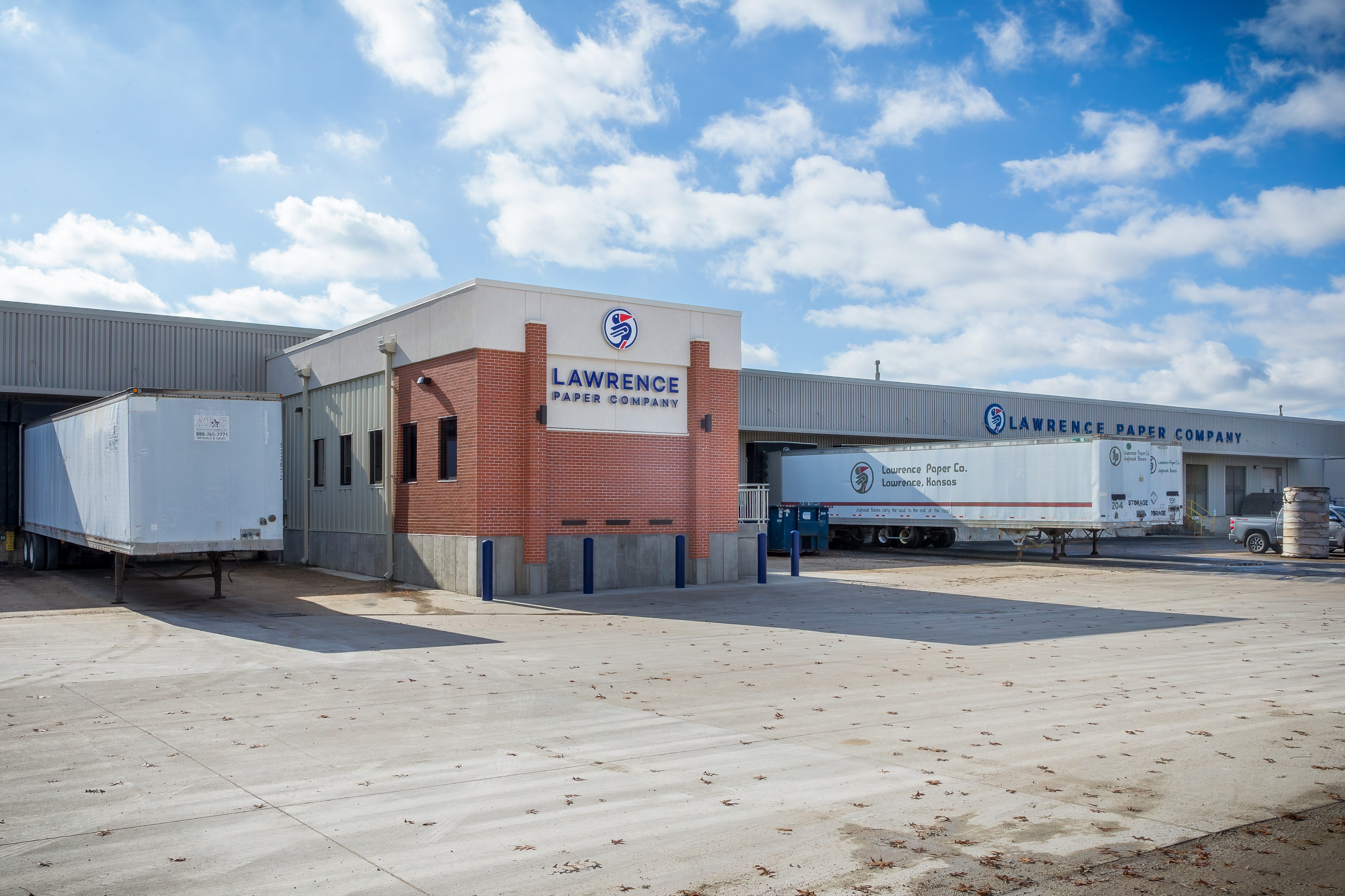 Lawrence Paper Company building with brick and white exterior, two semi-trailers parked nearby, and a blue sky with scattered clouds in the background.