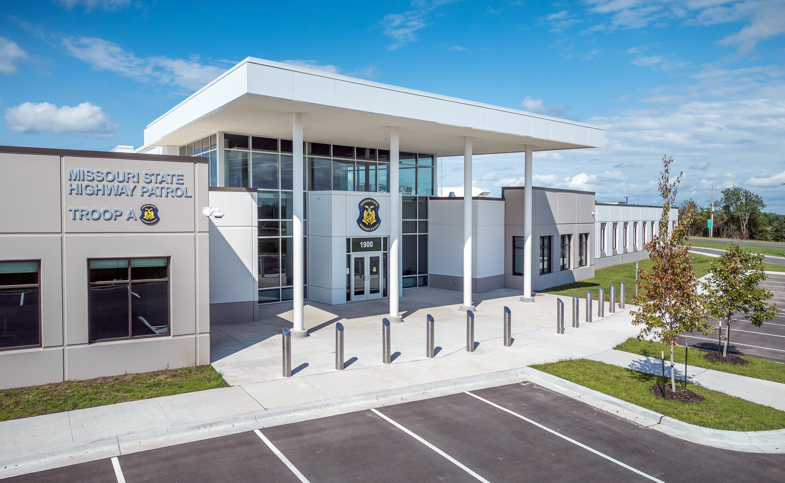 Modern building with large windows and pillars, labeled "Missouri State Highway Patrol Troop A." The entrance has a badge emblem above the doors, and the area is surrounded by a parking lot and some small trees.