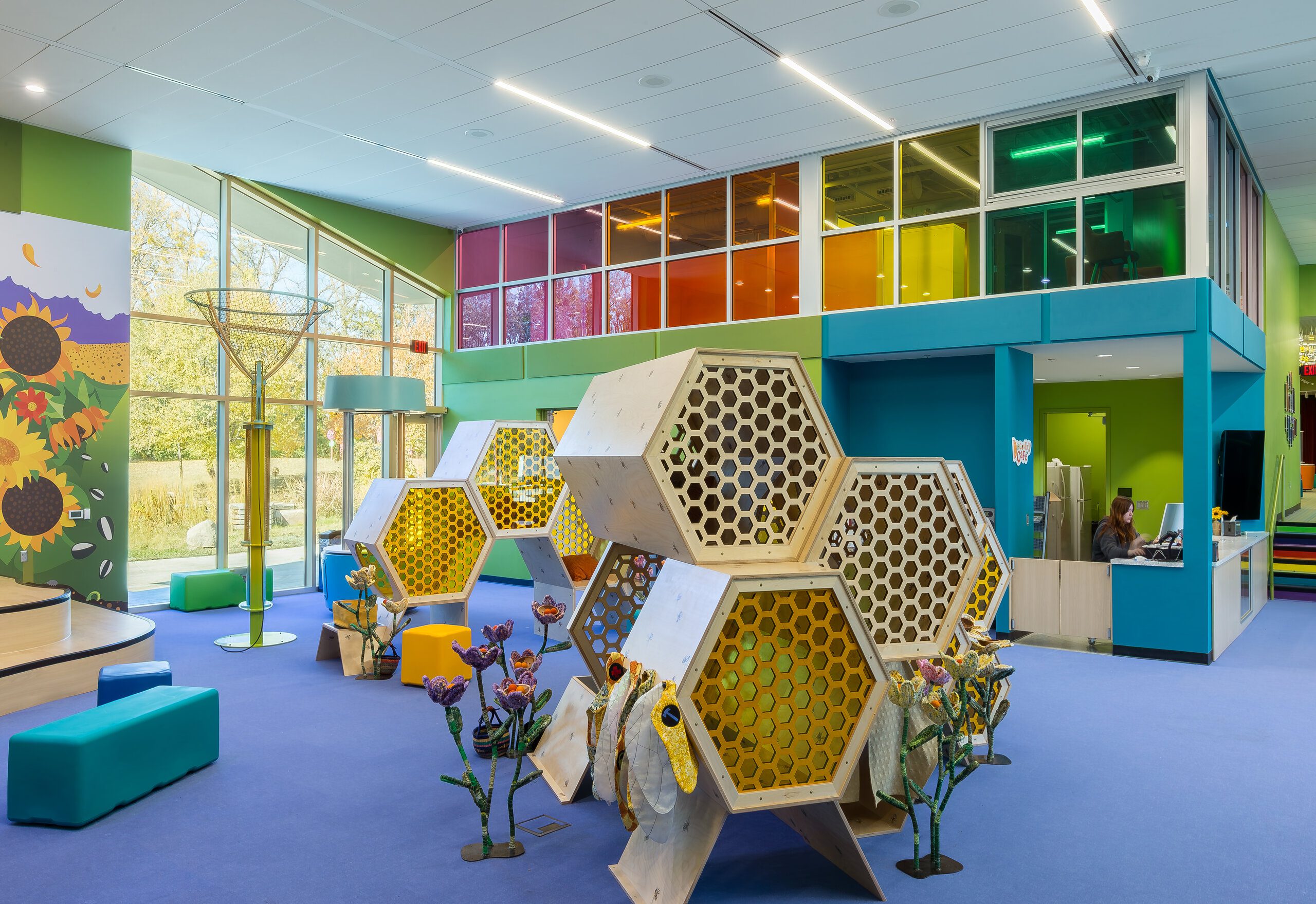 A bright, colorful indoor play area features large hexagonal honeycomb structures, flower decorations, soft seating, and a reception desk, with vibrant walls and natural light from large windows.