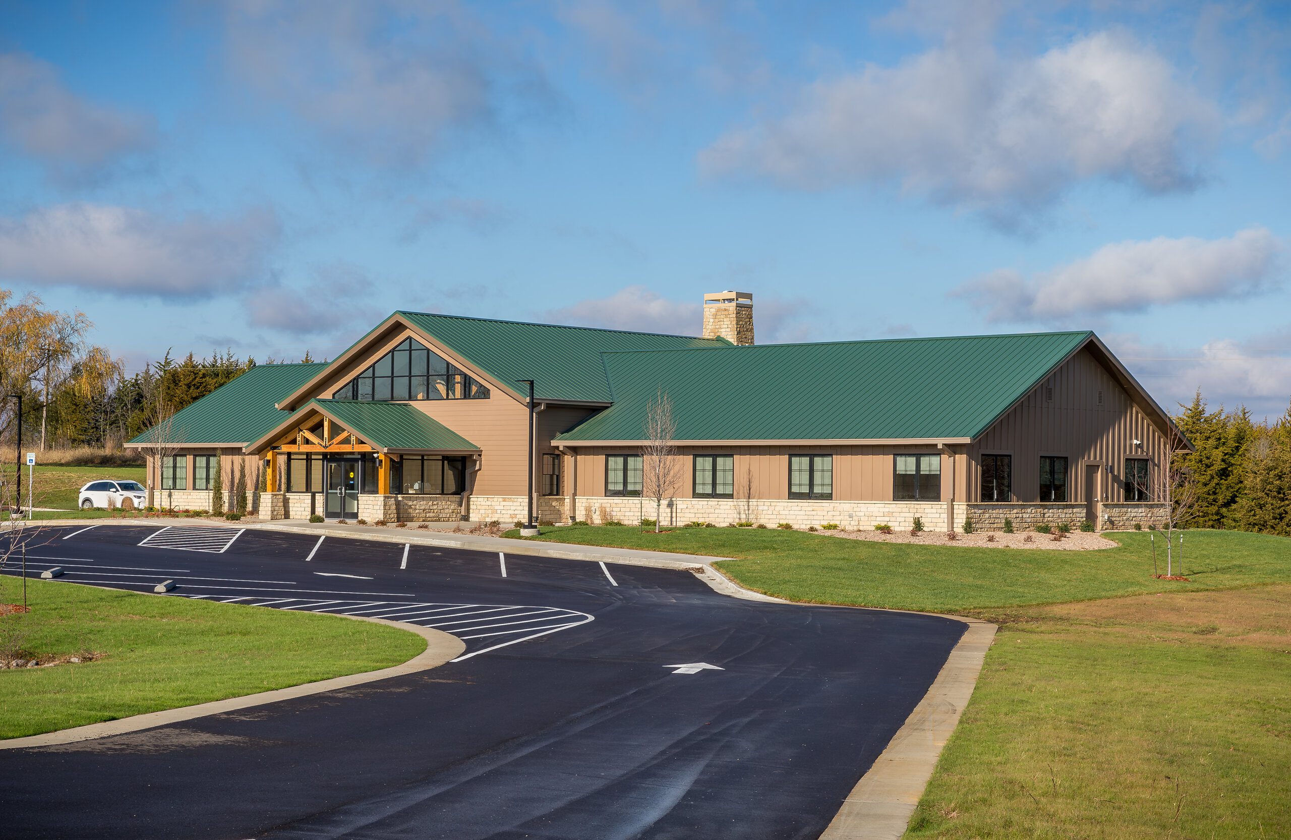 A one-story building with a green metal roof, large windows, and a wooden entrance sits next to an empty parking lot, surrounded by grass and trees under a partly cloudy sky.