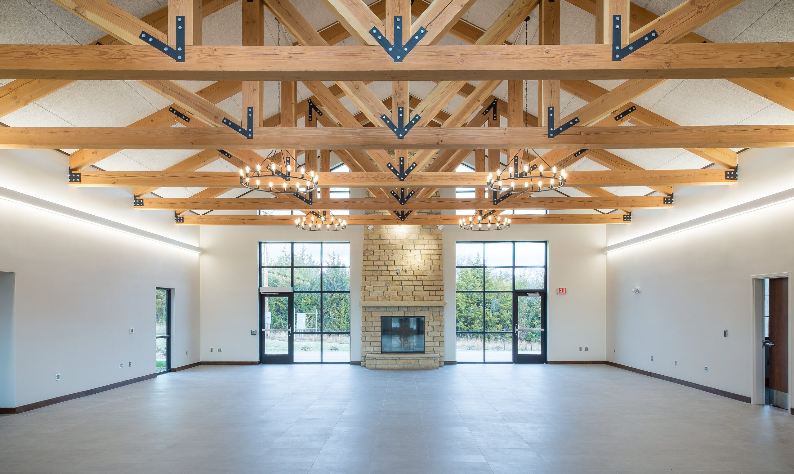 A spacious, empty hall with high wooden beam ceilings, large windows, a stone fireplace in the center, and modern chandeliers hanging from the ceiling. Natural light fills the room.