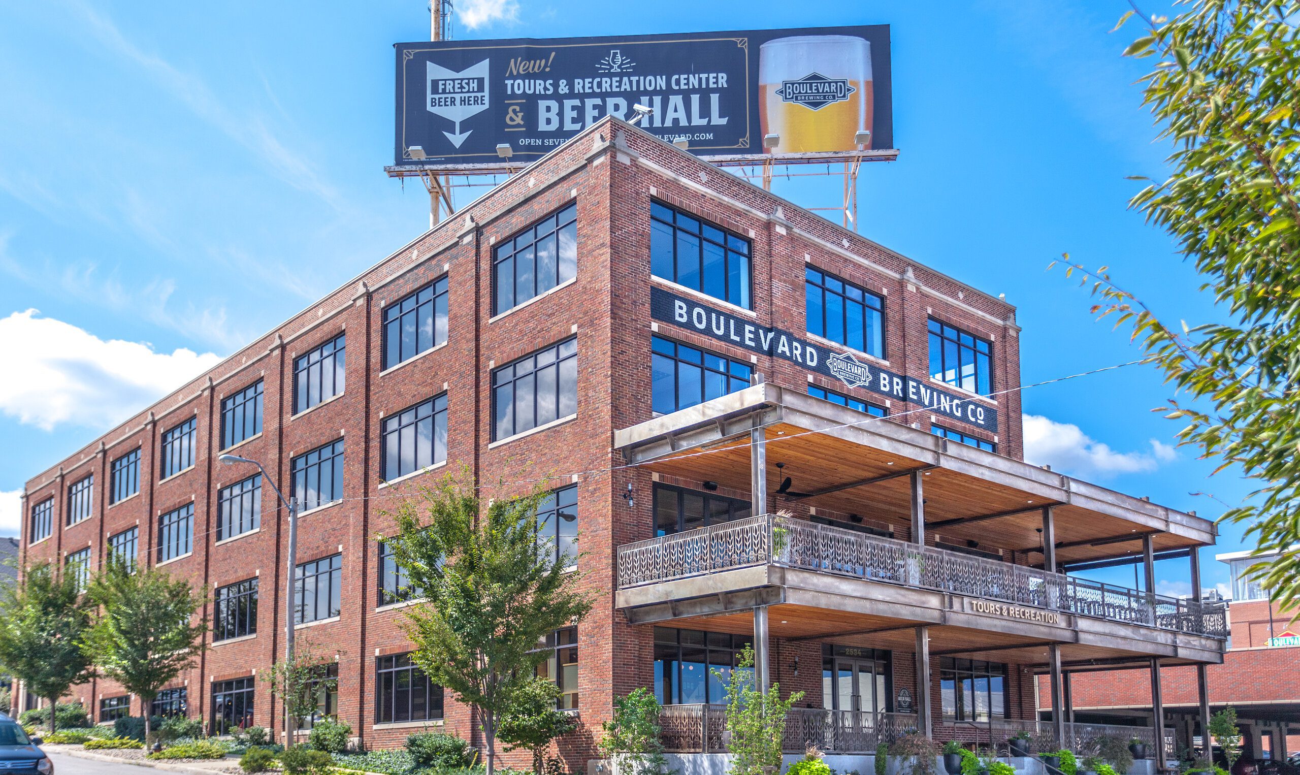 A large red brick building with “Boulevard Brewing Co.” signage and a covered balcony, surrounded by trees. A billboard on the roof advertises fresh beer, tours, a recreation center, and a beer hall.