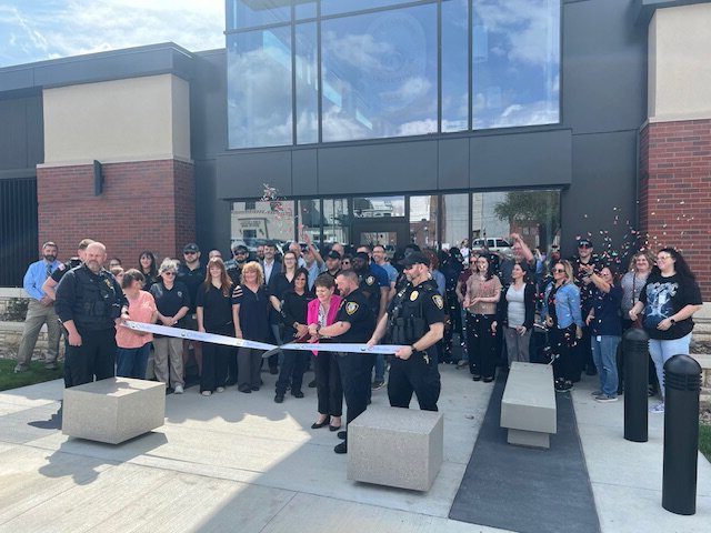 A large group of people, including police officers and officials, gather outside a modern building for a ribbon-cutting ceremony. Two women cut the ribbon while confetti is thrown in celebration.
