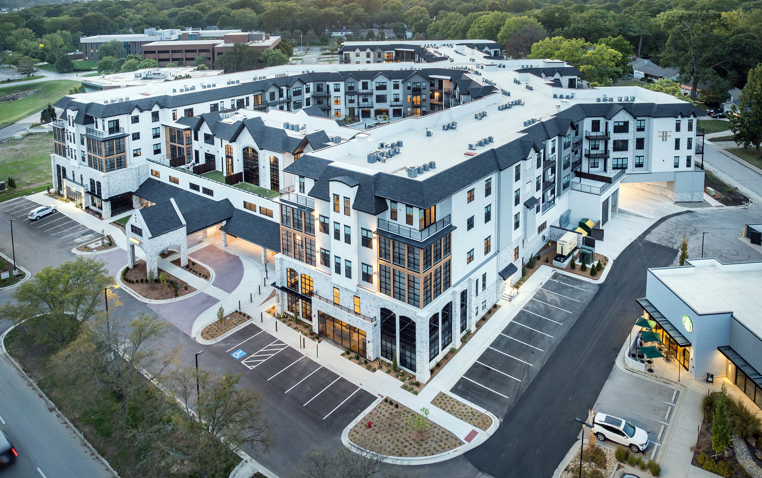 Aerial view of a large, modern apartment complex with white and gray exterior, multiple stories, and balconies, surrounded by parking lots, trees, and nearby buildings.