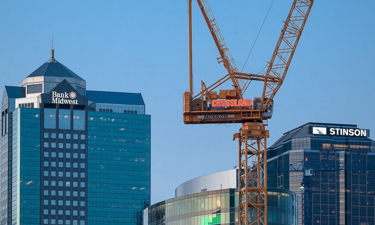 A construction crane stands in front of modern office buildings with "Bank Midwest" and "Stinson" logos visible on the tall glass towers against a clear sky.