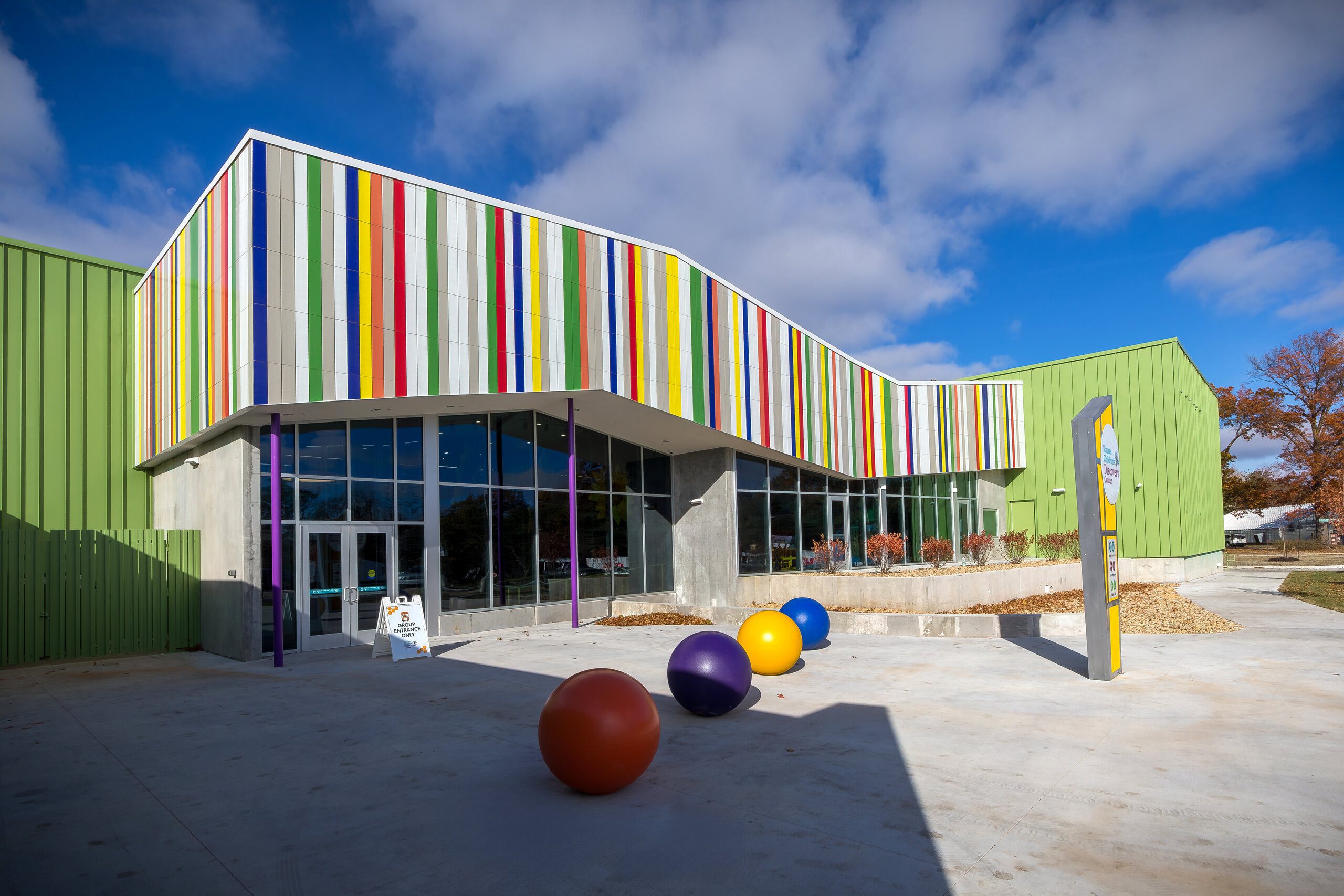 A modern building with green walls and a facade decorated with vertical multicolored stripes. Large glass windows and colorful spheres decorate the entrance under a partly cloudy sky.