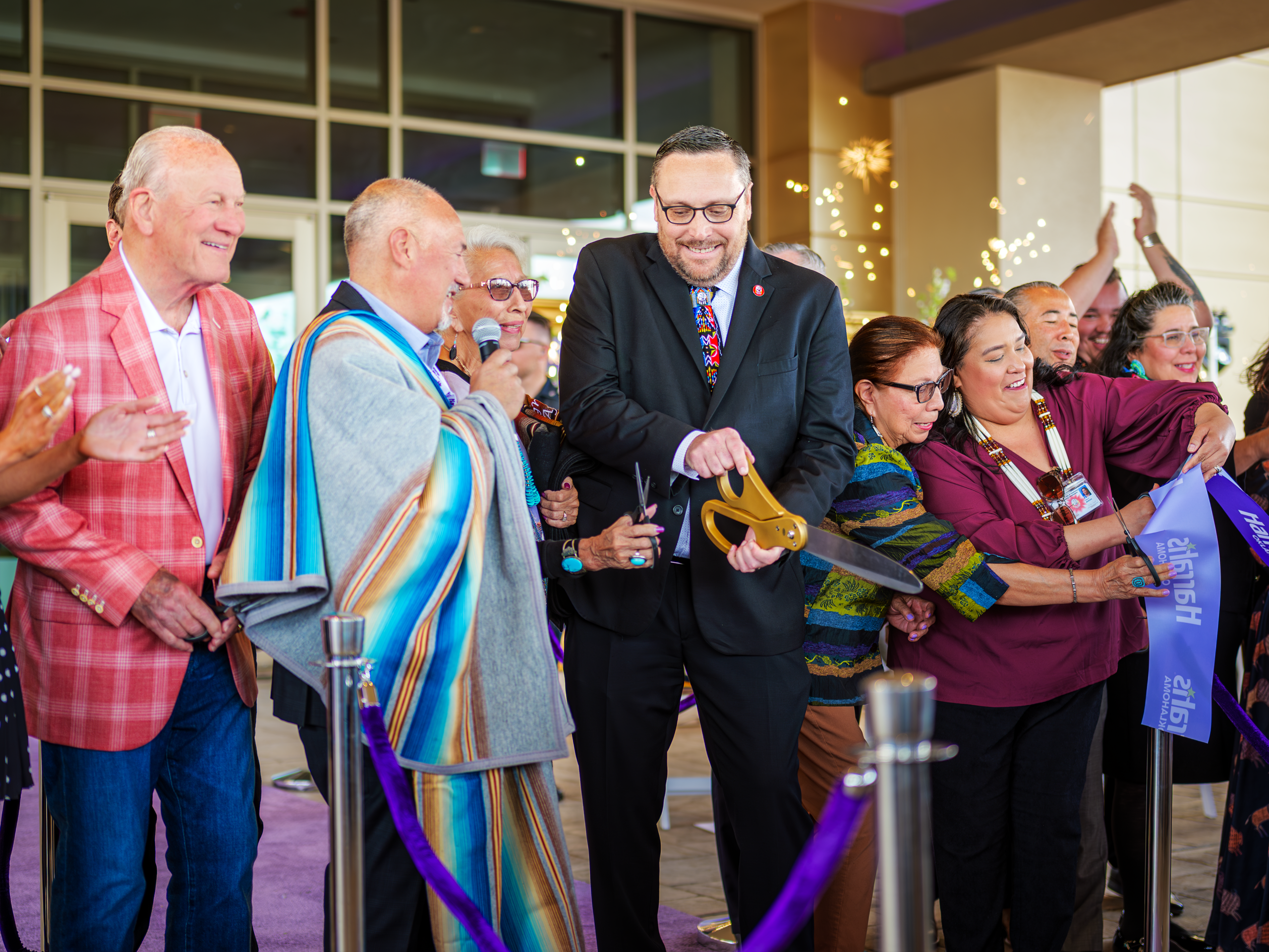 A group of people smile and cheer as a man in a suit uses large gold scissors to cut a purple ribbon at a grand opening event, surrounded by sparkling decorations and lively participants.