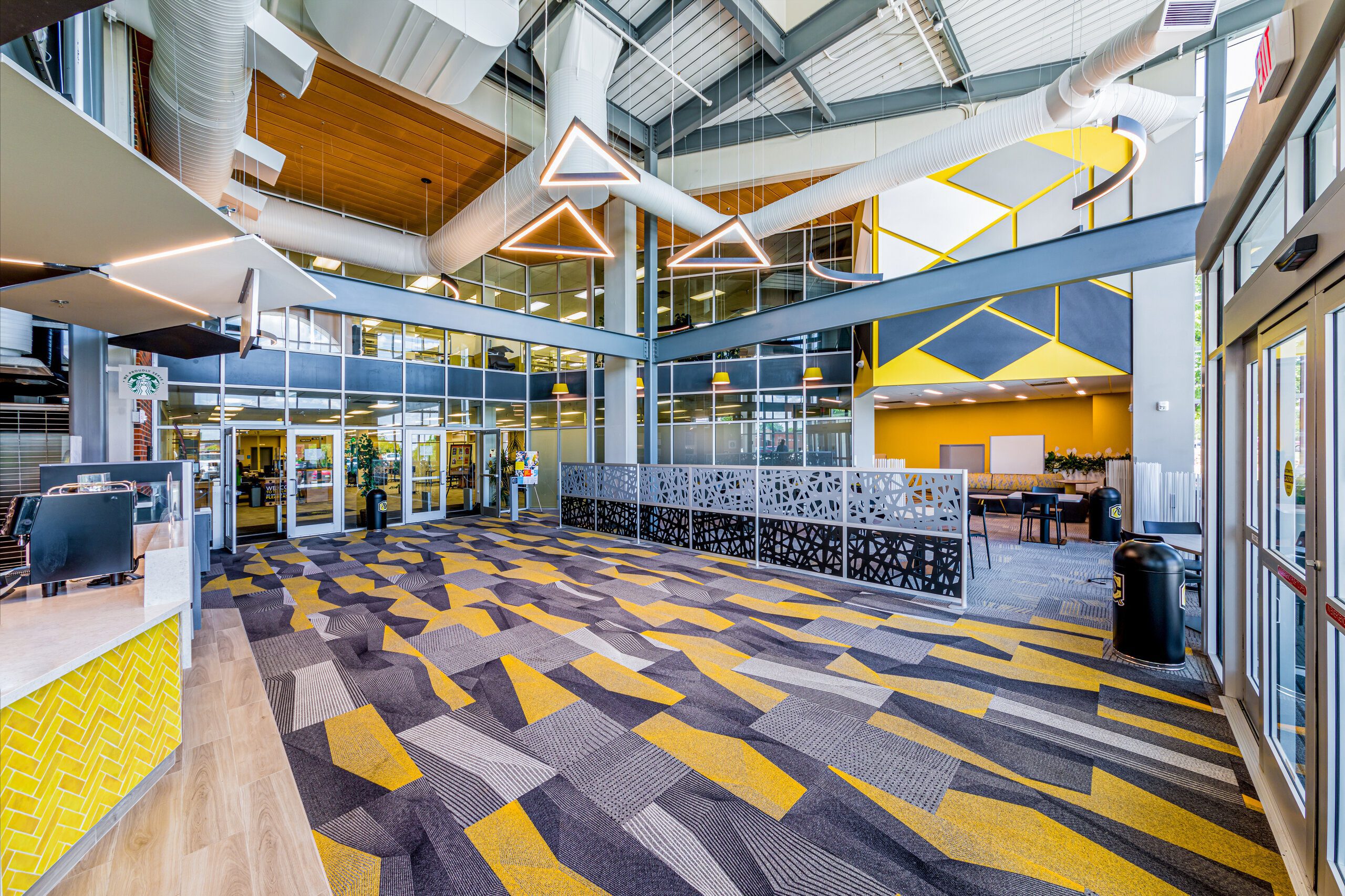 Spacious modern lobby with geometric carpet in gray and yellow tones, large windows, unique triangular ceiling lights, and a partial mezzanine with decorative railing. Reception desk and seating areas are visible.