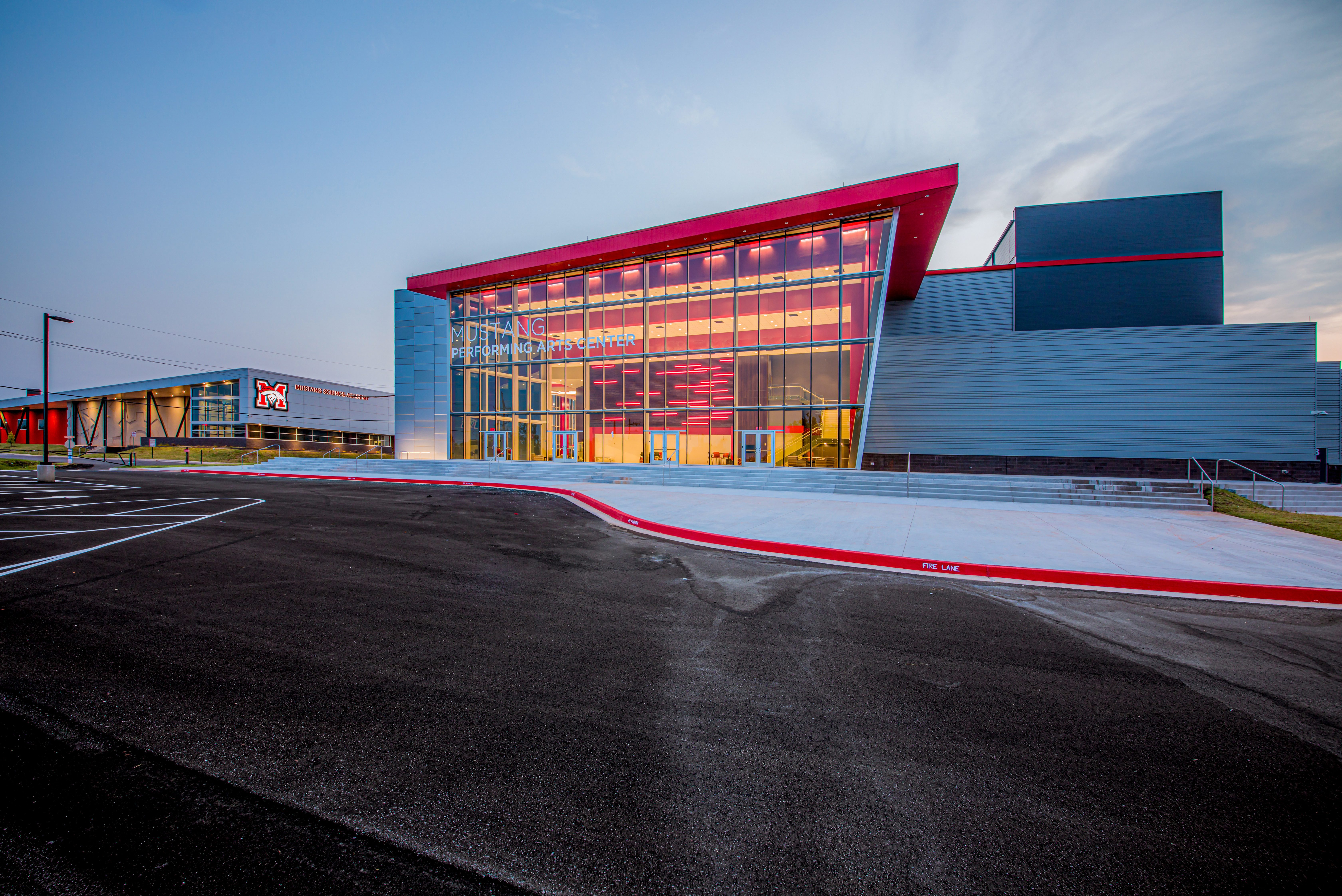 A modern building with large glass windows and red accents stands at dusk, featuring an empty parking lot and a wide sidewalk in front. The architecture is sleek and contemporary.
