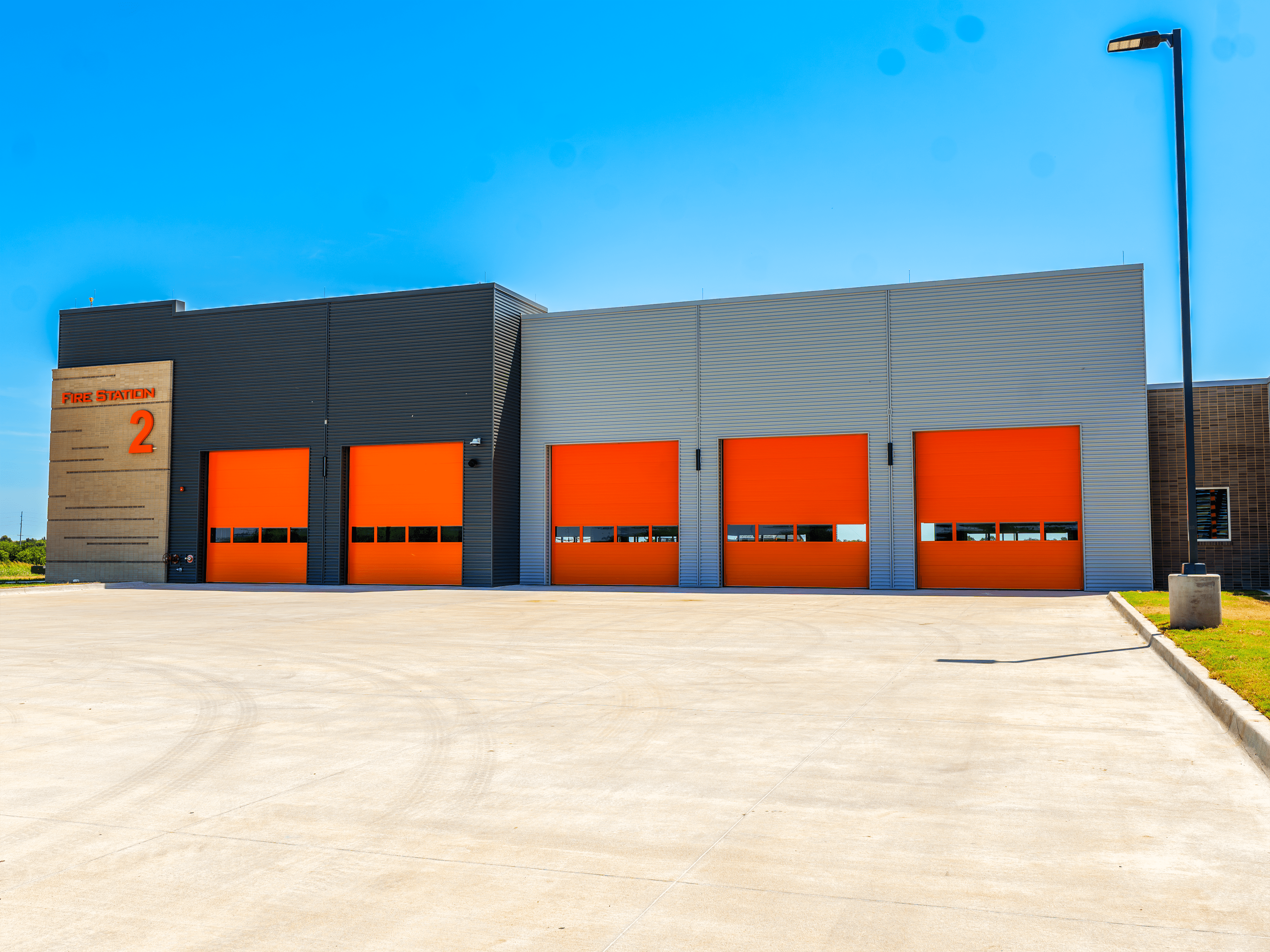 A modern fire station with five bright orange garage doors and a sign that reads "Fire Station 2" on a sunny day, with a large concrete driveway in front.
