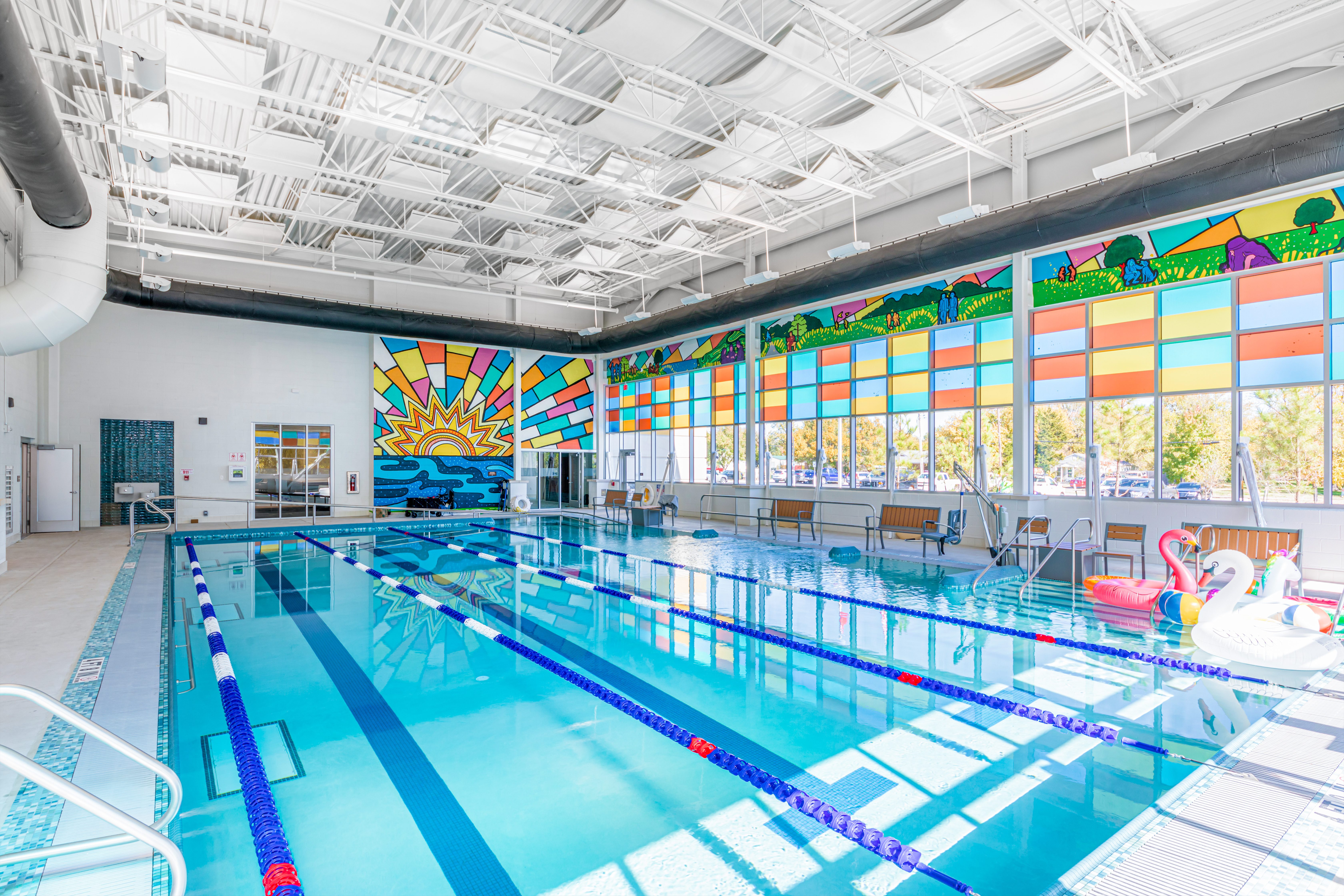 Indoor swimming pool with swim lanes, bright stained glass windows, a mural of a sunrise on the far wall, and inflatable pool toys. Sunlight streams through the colorful windows, illuminating the space.