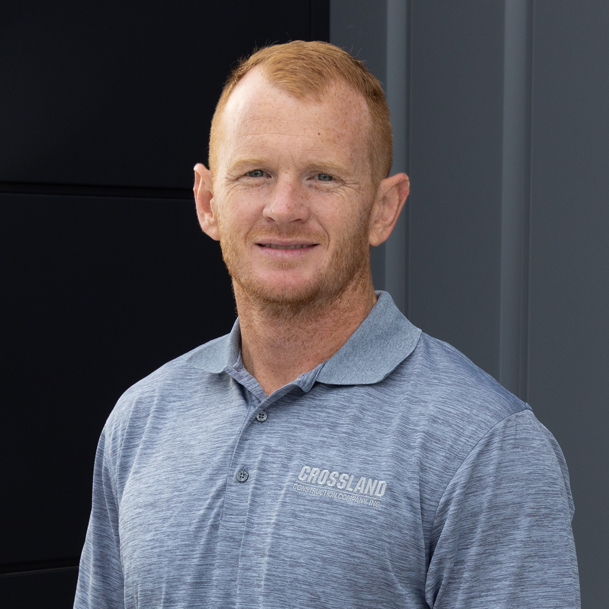 A man with short red hair wearing a light blue “Crossland” polo shirt stands in front of a dark and light gray background, smiling slightly at the camera.