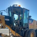 A construction worker wearing a safety vest and sunglasses operates a CAT 140M2 machine, sitting in the cab with sunlight shining behind him. The machine is on a dirt worksite under a clear sky.