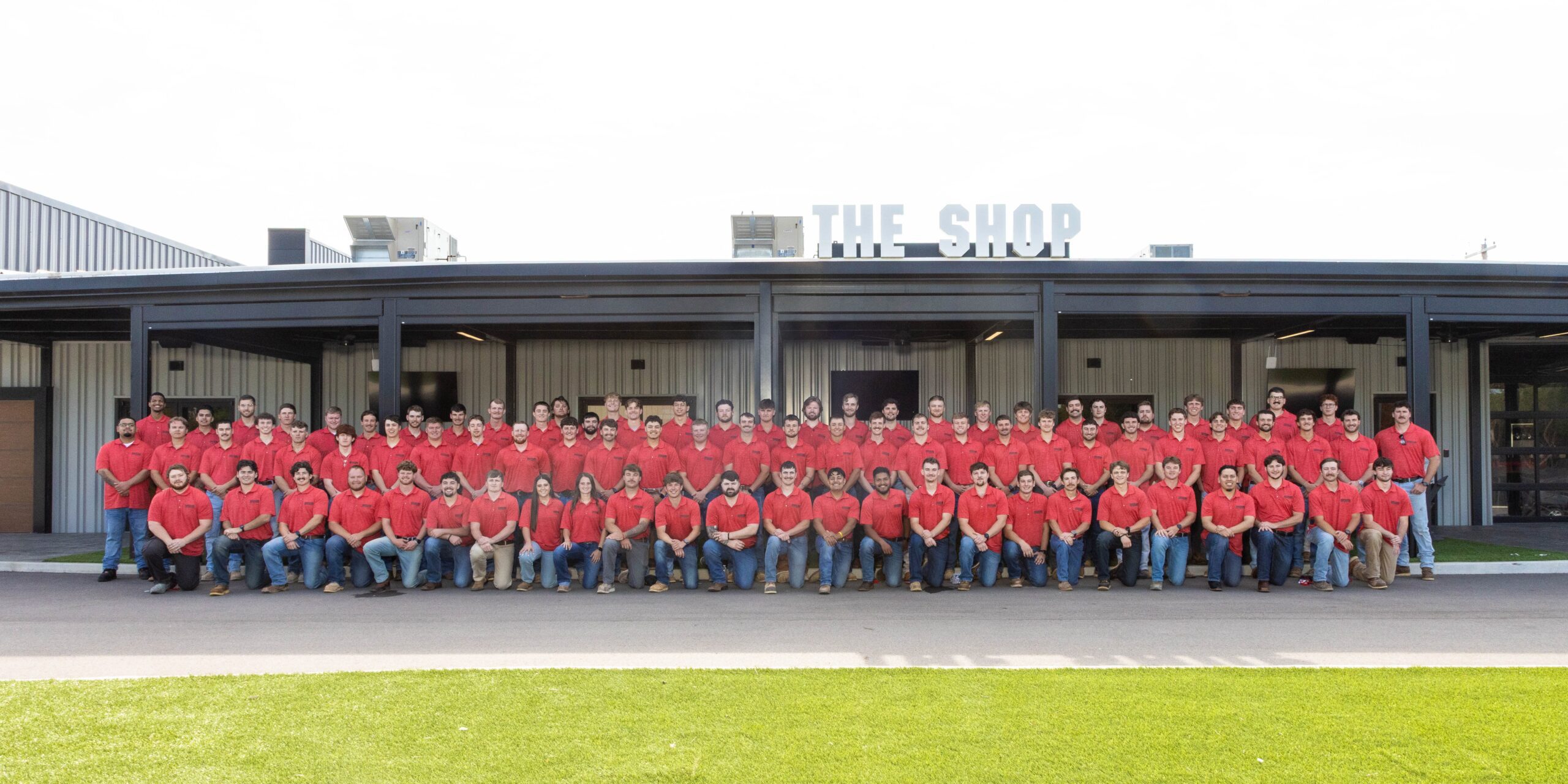 A large group of people wearing matching red shirts and jeans pose for a group photo in front of a building labeled “THE SHOP.” The group is arranged in rows on a paved area with green grass in the foreground.