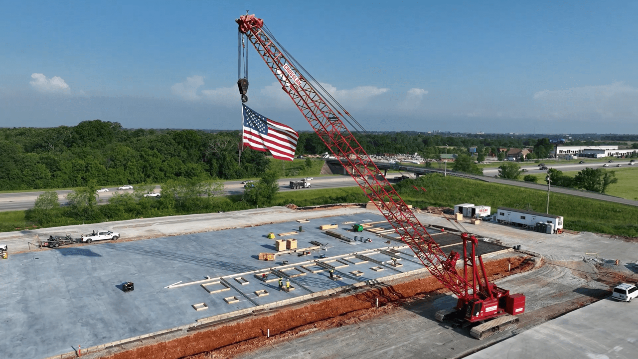 A large red construction crane with an American flag hanging from its hook sits on a building site with concrete foundation, surrounded by greenery and a highway under a clear blue sky.