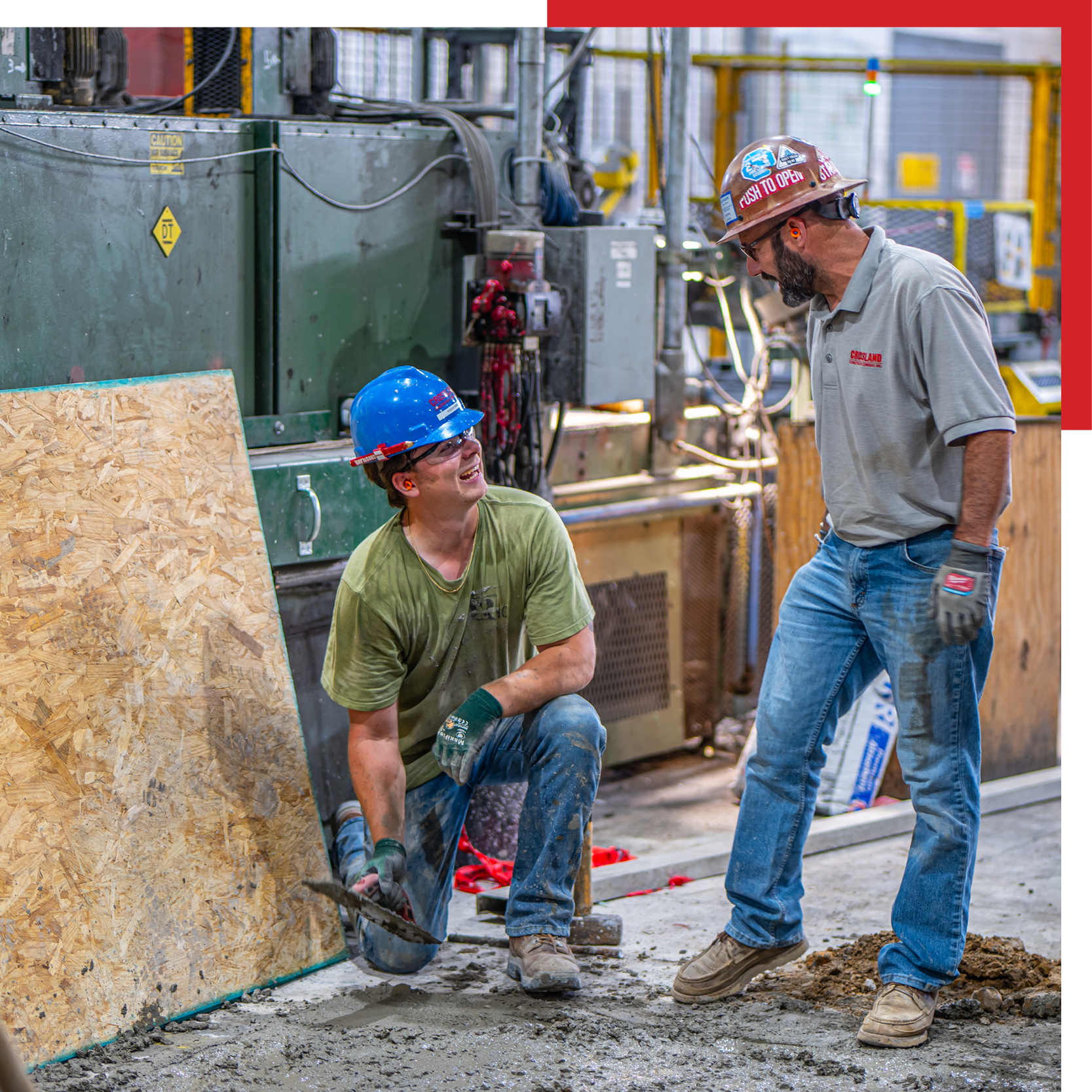 Two construction workers wearing hard hats and gloves are working with wet concrete indoors, near industrial equipment. One is kneeling and using a trowel, while the other stands talking to him.
